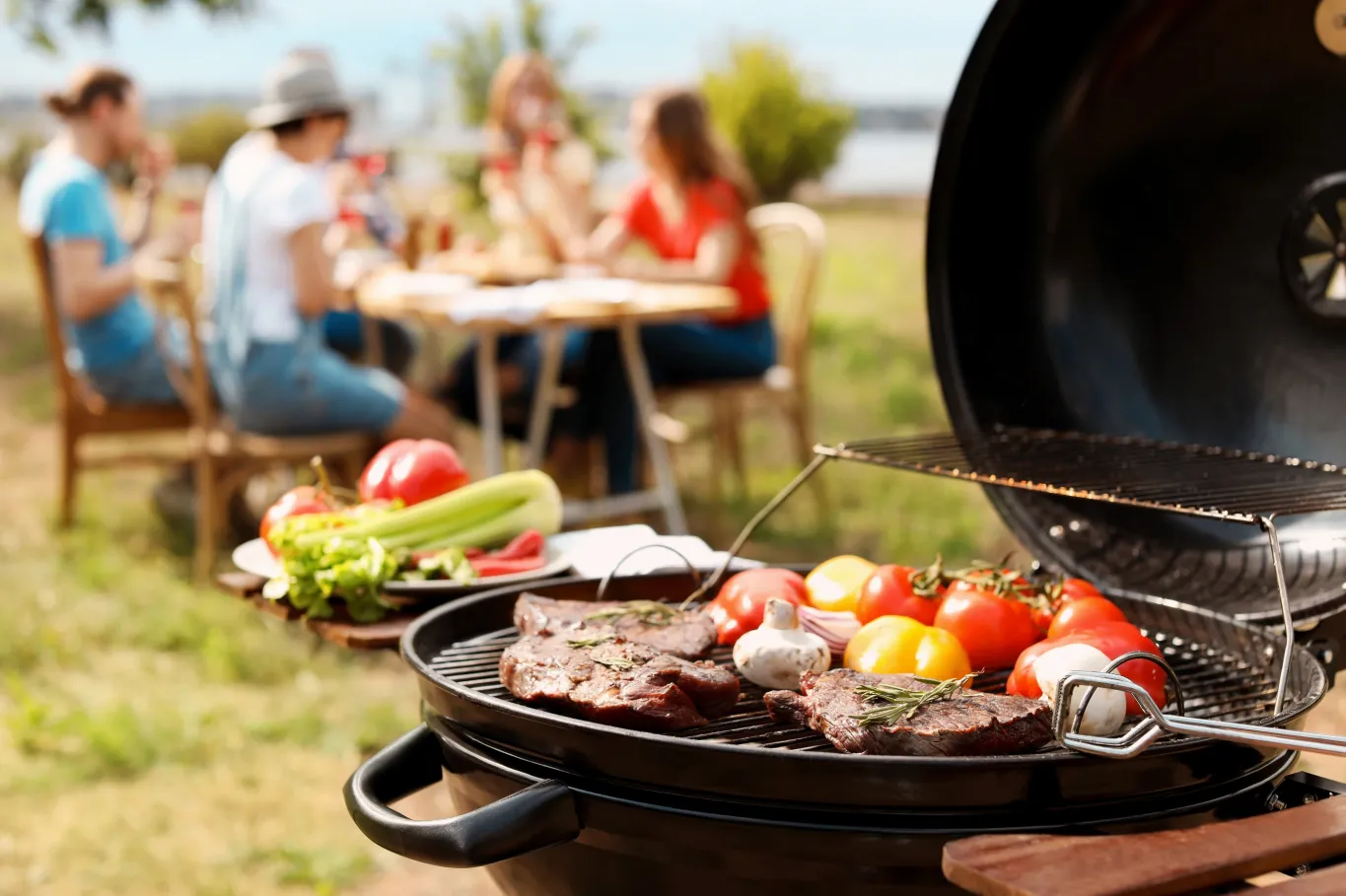 Photo montrant un gril dehors, en été, où cuisent des morceaux de viande et des légumes. À l’arrière-plan, des personnes sont assises autour d’une table.