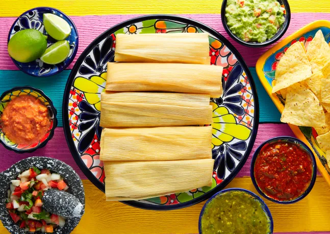 Photo d’une assiette de tamales entourée de sauces, de quartiers de limes, d’un guacamole, de croustilles de maïs.