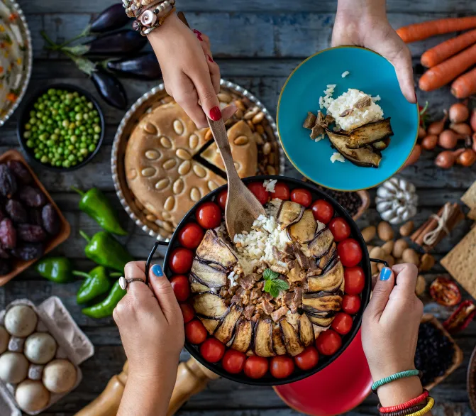 Photo montrant deux paires de mains tenant des plats de nourriture, et une table contenant différents plats et légumes.
